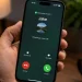 دست یک فرد آیفونی را در دست گرفته که صفحه‌ی تماس ورودی واتس‌اپ با نام Jake، عکس پروفایل کاربر، دکمه‌های قرمز Decline و سبز Accept و گزینه‌های Remind Me و Message را روی میز چوبی با لیوان قهوه و گلدان در پس‌زمینه نشان می‌دهد.