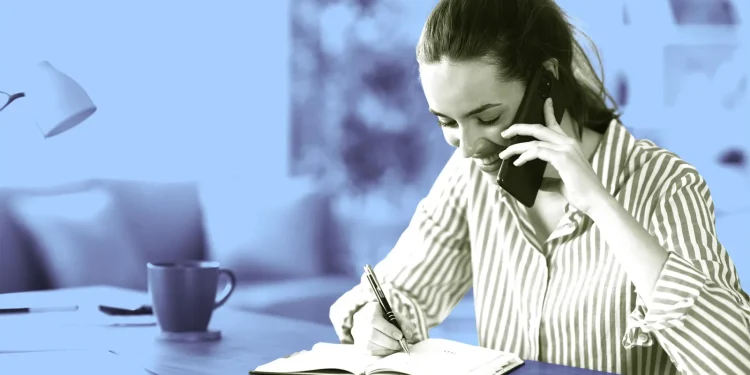 زن جوان در حال صحبت با گوشی موبایل و یادداشت‌برداری با خودکار در دفترچه روی میز کار
