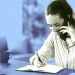 زن جوان در حال صحبت با گوشی موبایل و یادداشت‌برداری با خودکار در دفترچه روی میز کار