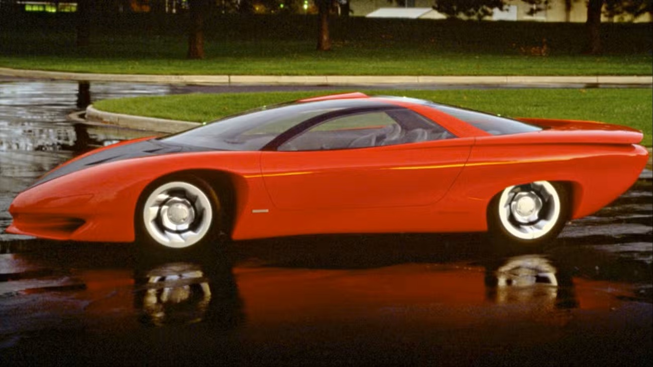 1988-pontiac-banshee-iv-concept ماشین اسپورت آیندهنگر قرمز که روی یک پیادهروی خیس با انعکاس، چمن سبز و درختان در پسزمینه پارک شده است.