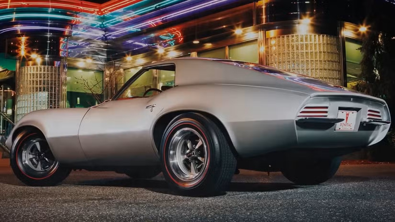 the- Greatest-pontiac-ever-made-11-7-screenshot-1 نمای سه چهارم عقب خودروی اسپرت پونتیاک کلاسیک نقرهای با چرخهای کرومی در شب، زیر چراغهای نئون.