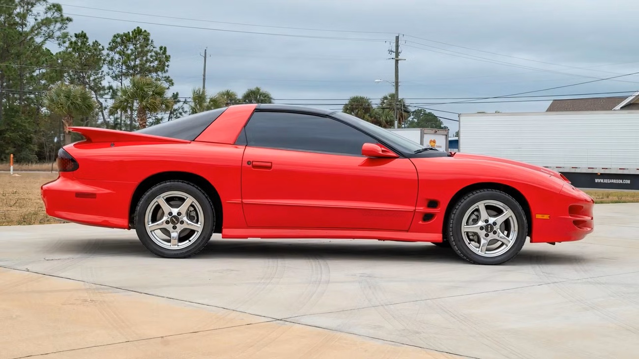 2001-pontiac-firebird-4 نمای جانبی یک ماشین اسپورت قرمز روشن که روی یک راهروی بتونی سبک پارک شده، شیشههای رنگی و یک اسپویلر در عقب قابل مشاهده است.