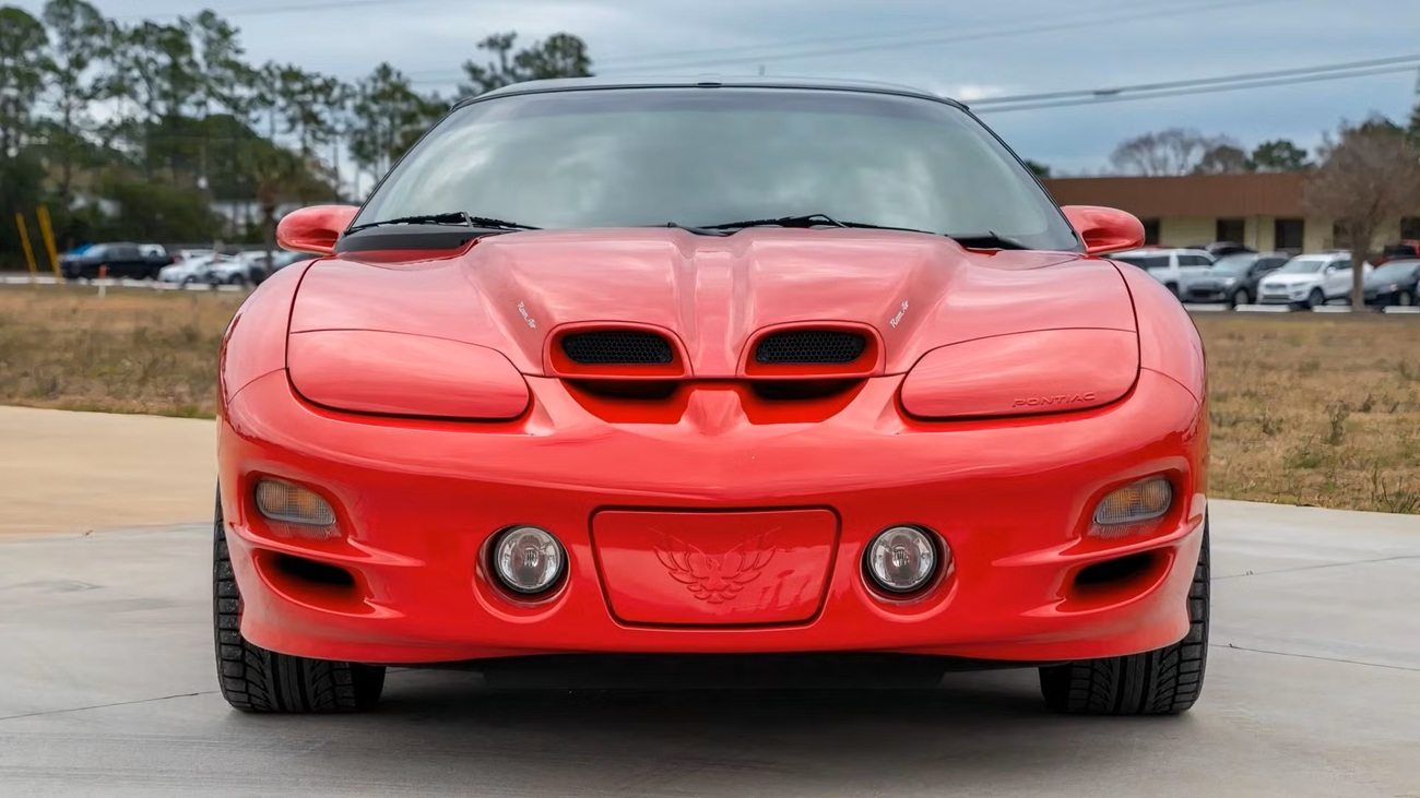 2001-pontiac-firebird-2 نمای جلوی یک خودروی اسپرت پونتیاک قرمز با اسکوپهای کاپوت دوگانه و چراغهای جلوی گرد، پارک شده روی سطحی بتونی با یک منطقه چمنزار در پسزمینه.