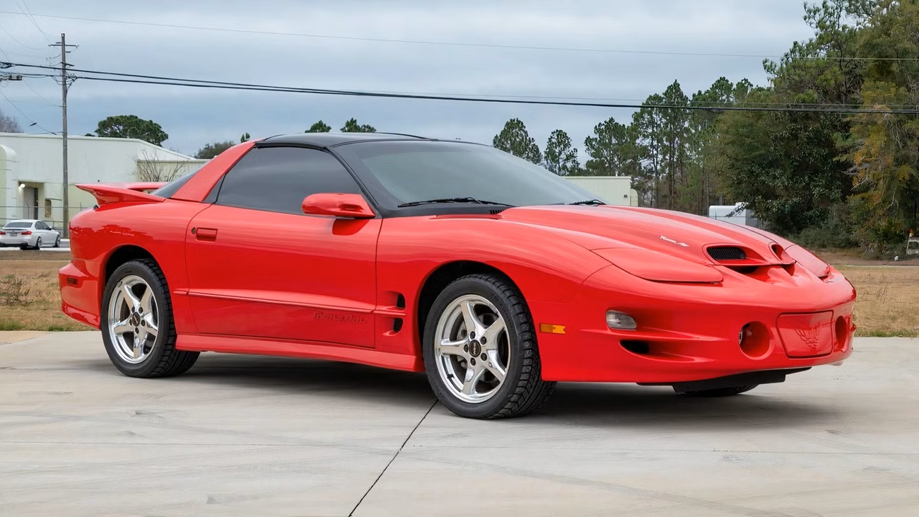 2000-pontiac-firebird-1 کوپه اسپرت Pontiac Firebird Trans Am قرمز با اسپویلر، پارک شده در یک راهروی سیمانی با شیشههای رنگی.