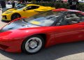 Red Ferrari LaFerrari with black hood and silver wheels, parked at a car show with a yellow sports car in the background and people strolling nearby.