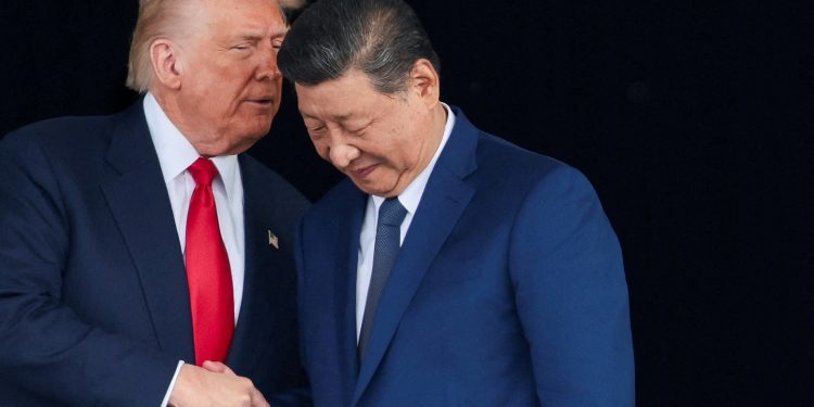 Two men in dark suits shake hands and lean together against a dark background, one wearing a red tie and the other a blue tie, as they converse closely.