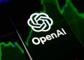 گوشی هوشمند با لوگوی سفید و نام OpenAI روی صفحه نمایش در مقابل پس‌زمینه سبز و تیره نمودارهای بازارهای مالی.