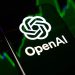 گوشی هوشمند با لوگوی سفید و نام OpenAI روی صفحه نمایش در مقابل پس‌زمینه سبز و تیره نمودارهای بازارهای مالی.