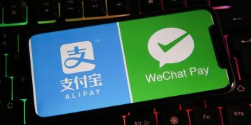 یک گوشی هوشمند روی صفحه‌کلید گیمینگ با نور پس‌زمینه رنگی که لوگوهای Alipay و WeChat Pay را روی نمایشگر خود نشان می‌دهد.