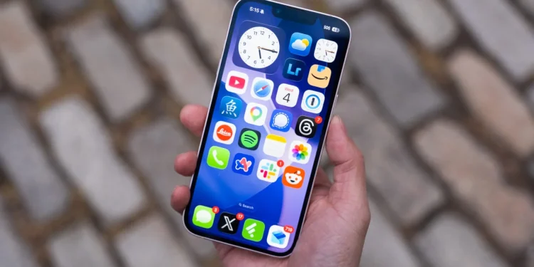 ویژگی جدید iOS 27 امکان استفاده از همان عدد را در دو آیفون فراهم می کند 1 Hand holding a smartphone showing a colorful app grid on the home screen over a cobblestone background.