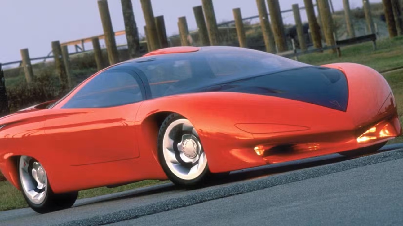 1988-pontiac-banshee-concept خودروی اسپورت آیندهنگر قرمز با شیشه جلویی به سبک سایهبان رنگی مشکی در جاده، پسزمینه شهری پشت حصار.