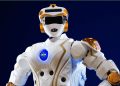NASA humanoid robot in white armor with orange joints and a visible NASA logo on the chest, posed against a dark blue background.