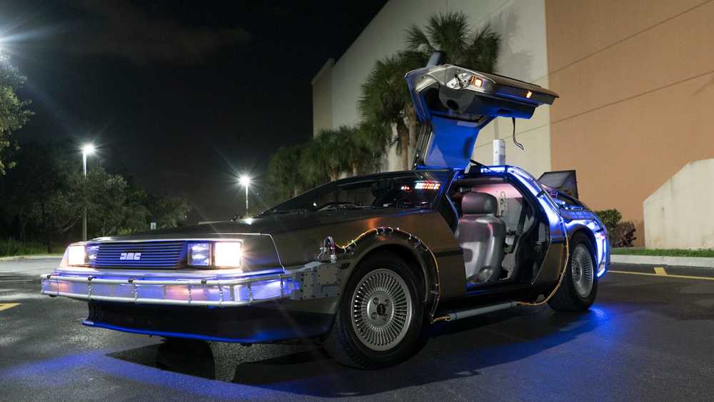 IMG_20261001_125715_490 ماشین زمان DeLorean DMC-12 با درهای باز بال مرغان، زیردرخشش آبی، پارک شده در پارکینگ شب تاریک.