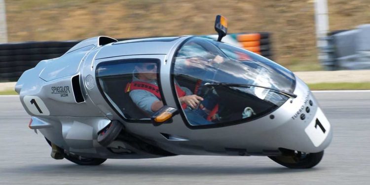 با Peraves EcoMobile آشنا شوید. ترکیبی از موتور سیکلت، هواپیما و ماشین با طراحی دیوانه کننده 1 Single-seat, silver enclosed race car tilting into a turn on a racetrack, driver visible inside wearing a red harness (car number 1).