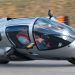 با Peraves EcoMobile آشنا شوید. ترکیبی از موتور سیکلت، هواپیما و ماشین با طراحی دیوانه کننده 8 Single-seat, silver enclosed race car tilting into a turn on a racetrack, driver visible inside wearing a red harness (car number 1).
