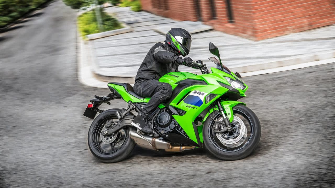 2027-kawasaki-ninja-650-19-16x9 موتورسوار با دنده مشکی که به یک کاوازاکی نینجا 650 سبز تیز در خیابان شهری منحنی با دیوار آجری تکیه داده است.