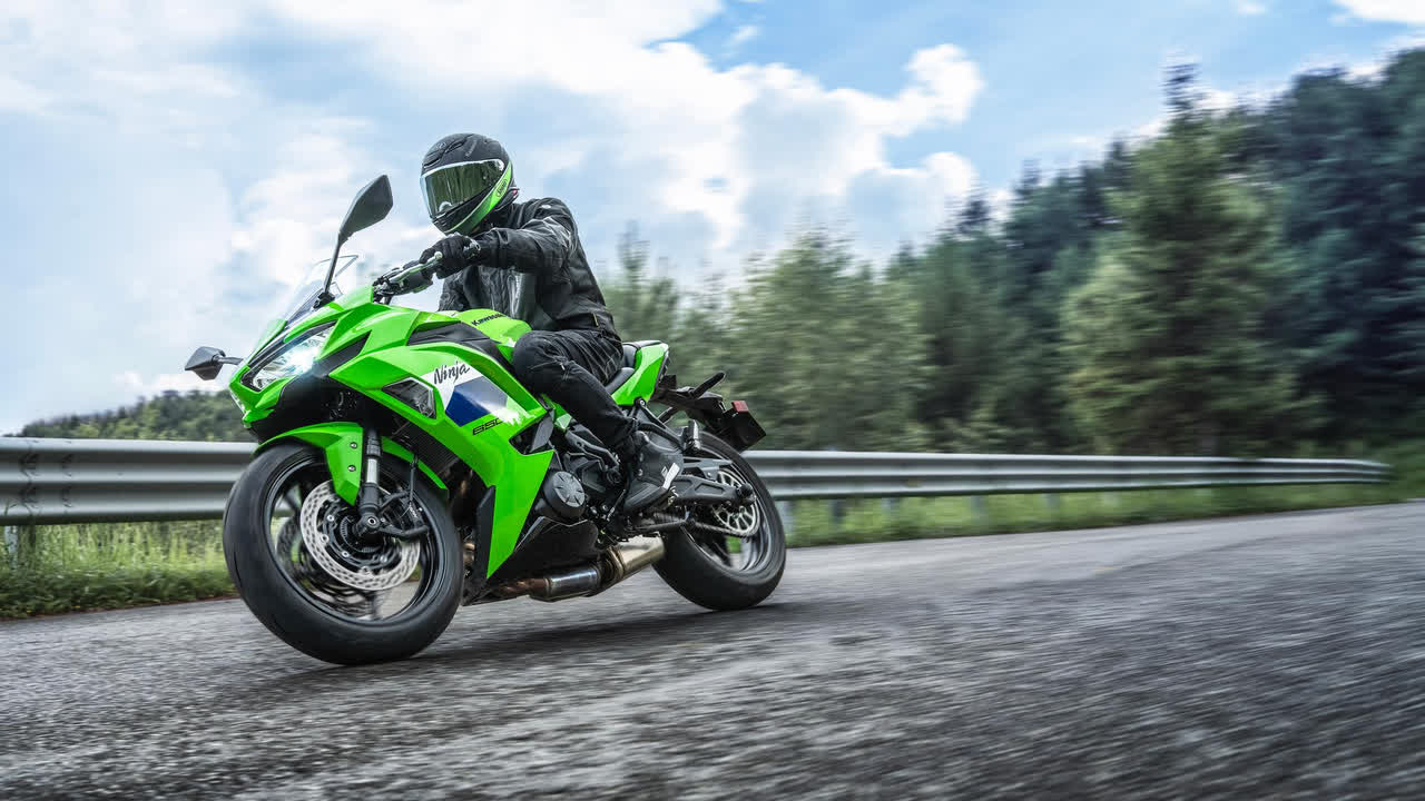 2027-kawasaki-ninja-650-15-16x9 موتورسوار با دنده مشکی روی یک دوچرخه اسپرت کاوازاکی سبز رنگ نئون در جاده ای با نرده محافظ و درختان در پس زمینه به چرخشی تکیه می دهد.