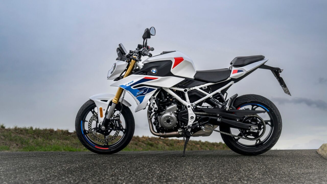 موتورسیکلت BMW سفید با برچسب‌های آبی و قرمز، در نمای جانبی پارک شده در لبه جاده نشان داده شده است.