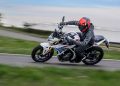 Motorcyclist in black gear and a red helmet leans into a sharp turn on a white sport bike along a road, with motion blur in the background.