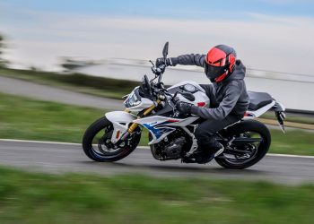 Motorcyclist in black gear and a red helmet leans into a sharp turn on a white sport bike along a road, with motion blur in the background.