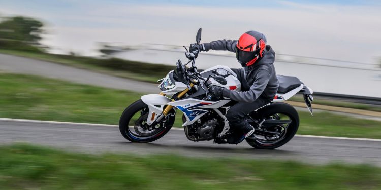Motorcyclist in black gear and a red helmet leans into a sharp turn on a white sport bike along a road, with motion blur in the background.