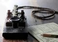 Vintage telegraph key with a coiled wire and a nearby instruction sheet on a dark wooden table, suggesting Morse code use
