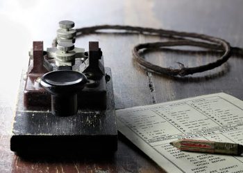 Vintage telegraph key with a coiled wire and a nearby instruction sheet on a dark wooden table, suggesting Morse code use