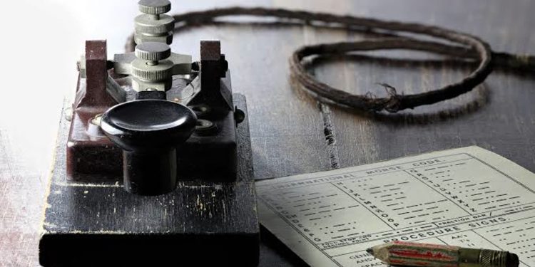 Vintage telegraph key with a coiled wire and a nearby instruction sheet on a dark wooden table, suggesting Morse code use