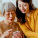 صدای شما ممکن است نشان دهد که چقدر سریع پیر می شوید 8 Two women, an elderly woman and a younger woman, look at a smartphone together.
