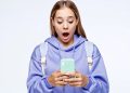Young woman in a lavender hoodie and backpack looks surprised while checking her mint green smartphone.