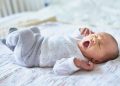 چرا هفته اول زندگی ممکن است برای سلامت آینده کودک مهم باشد؟ 17 Yawning newborn baby lying on a light blanket, wearing white pajamas with small stars.