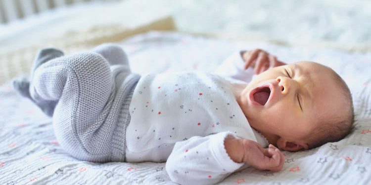 چرا هفته اول زندگی ممکن است برای سلامت آینده کودک مهم باشد؟ 1 Yawning newborn baby lying on a light blanket, wearing white pajamas with small stars.