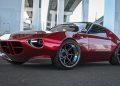 Low-angle view of a glossy red sports coupe with wide aftermarket wheels parked under an elevated highway pillar area in an urban setting.