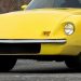 Front view of a bright yellow vintage sports car, showing round headlight, chrome bumper, and curved body lines on a pavement surface.