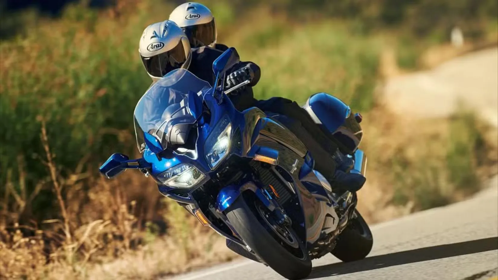 Two riders wearing helmets on a blue sport motorcycle leaning into a curve on a sunny rural road.