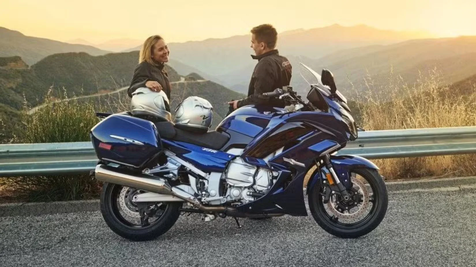 Two motorcyclists stand beside a blue sport motorcycle on a mountain road at sunset, wearing jackets with two helmets resting on the seat.