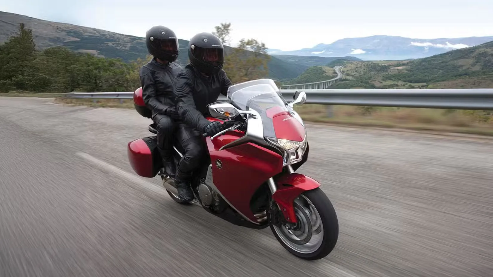 Two riders in black protective gear on a red touring motorcycle leaning into a curve on a winding mountain road.