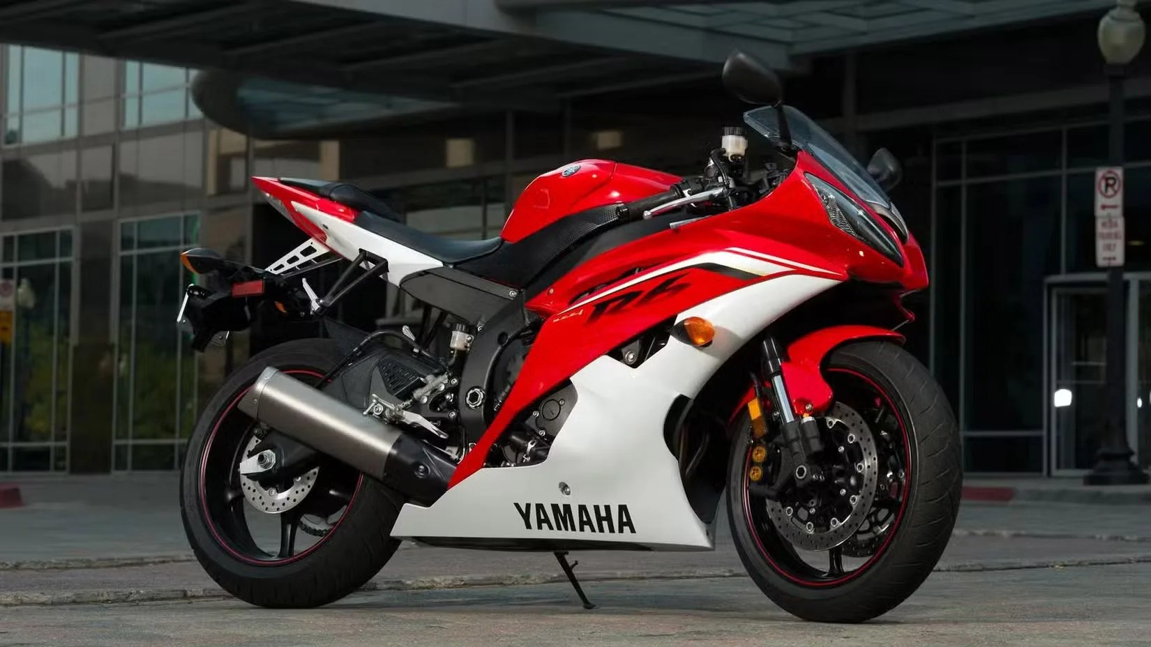 Red and white Yamaha sport motorcycle parked on a city street with a modern glass building in the background.