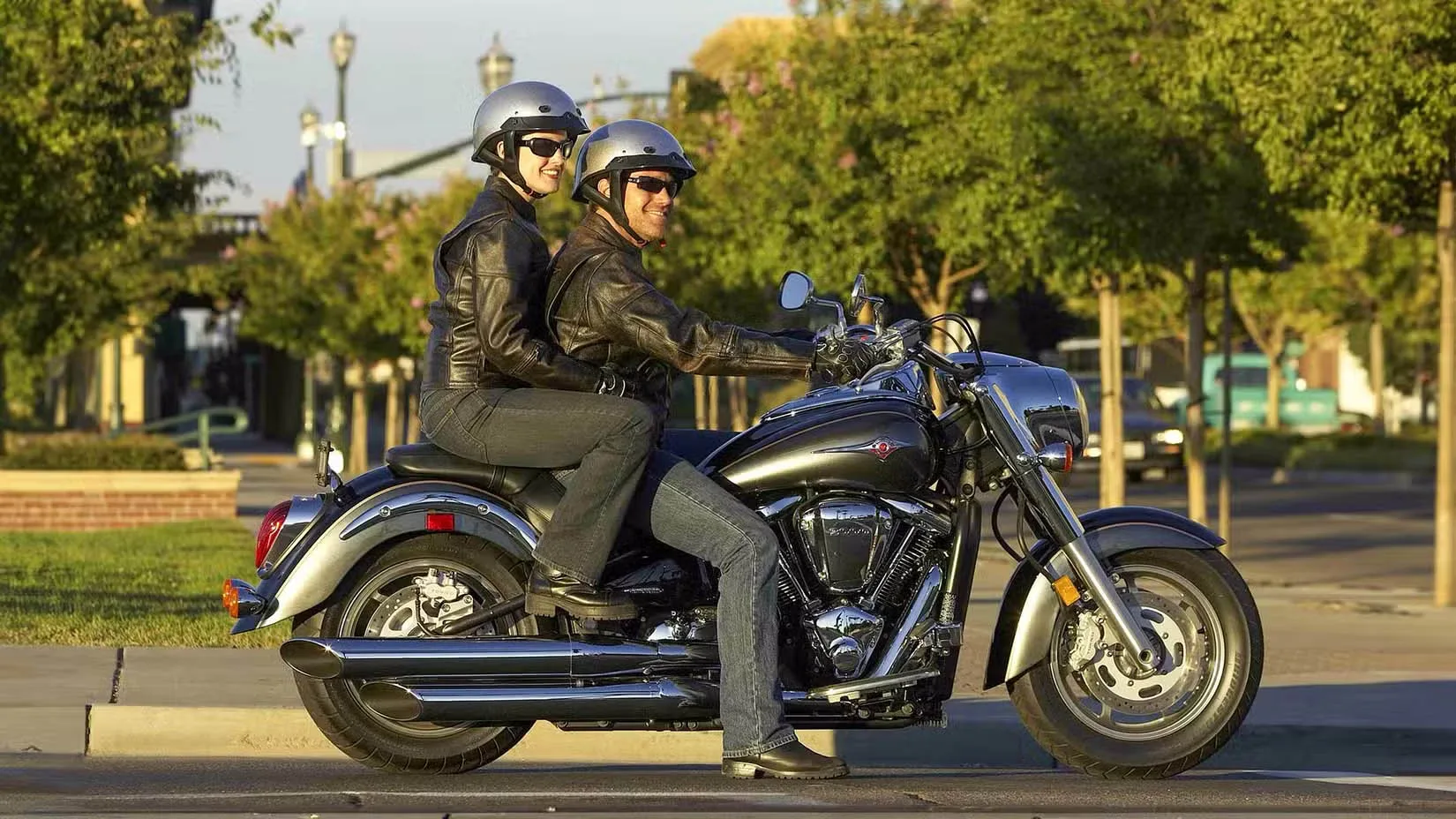 Couple riding a black touring motorcycle in leather jackets and helmets on a sunny street.