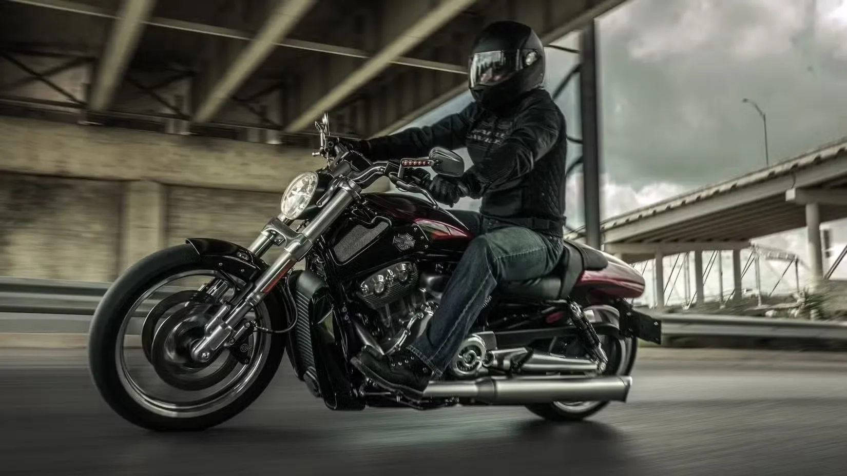 Motorcyclist in a black helmet and jacket leans into a turn on a red and black cruiser beneath an overpass.