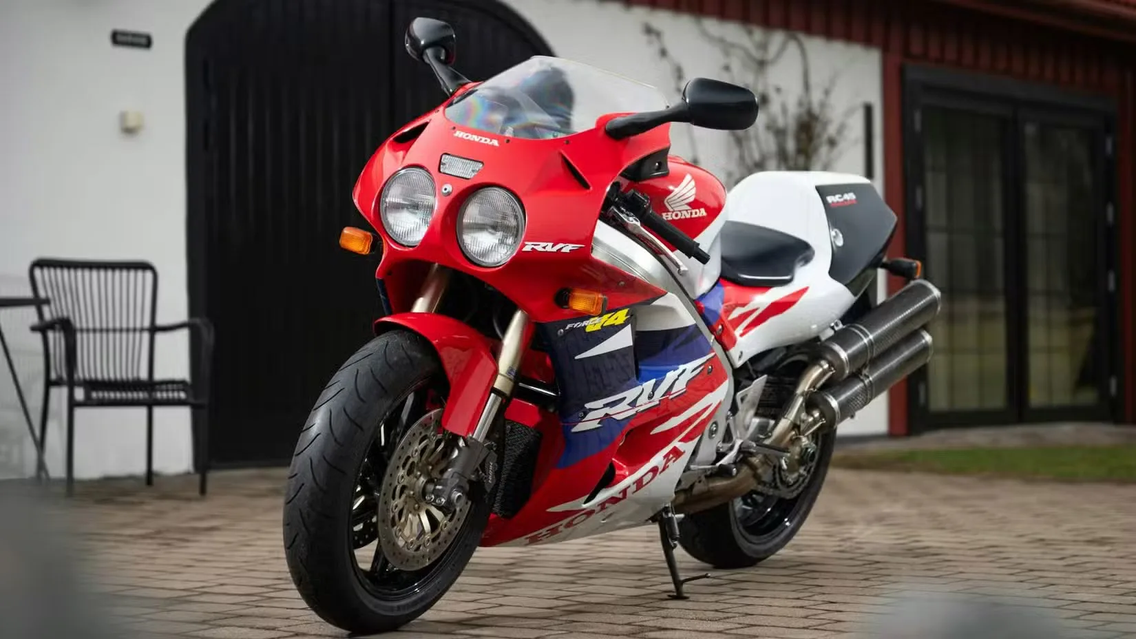 Red Honda sport motorcycle with dual round headlights and white-blue graphics, parked on a brick driveway in front of a house.