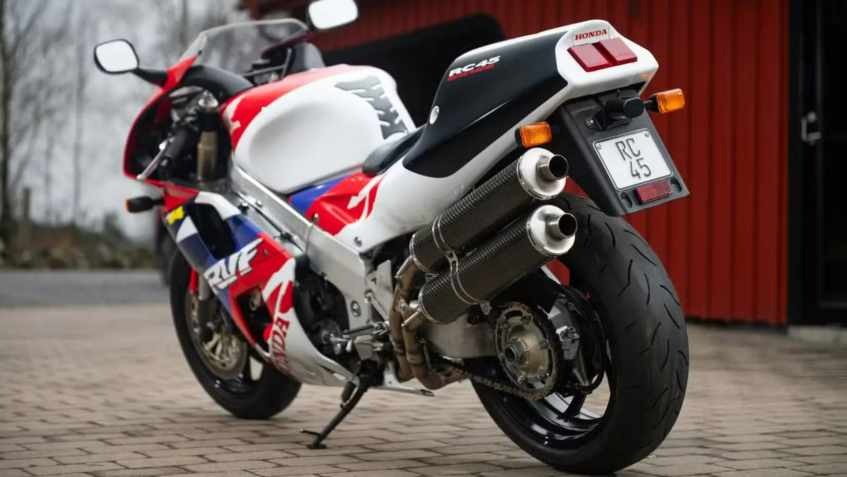 Sport motorcycle with red, white, and blue racing livery parked on a brick surface, showing dual exhausts and a Honda RC45 badge on the tail.
