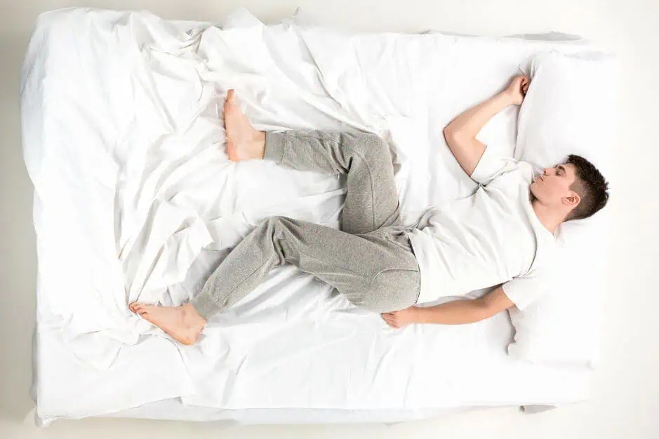 1000084729 Top-down view of a man lying on a white bed wearing a white T-shirt and gray joggers.