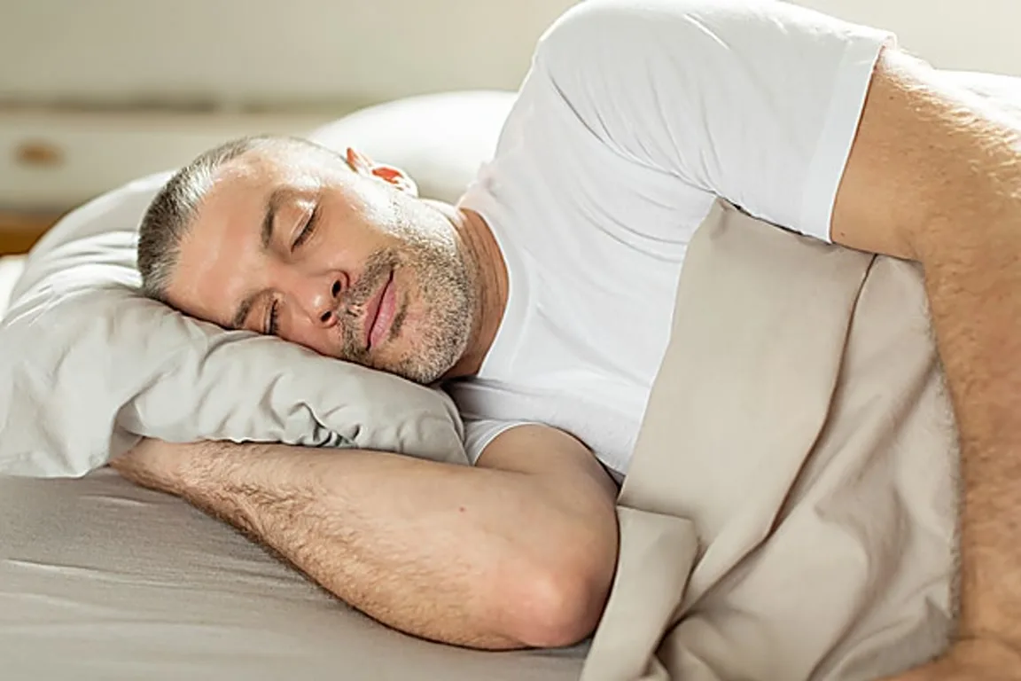 1000084733 Man sleeping on his side with a pillow under his head in a bed, wearing a white t‑shirt and beige blanket nearby