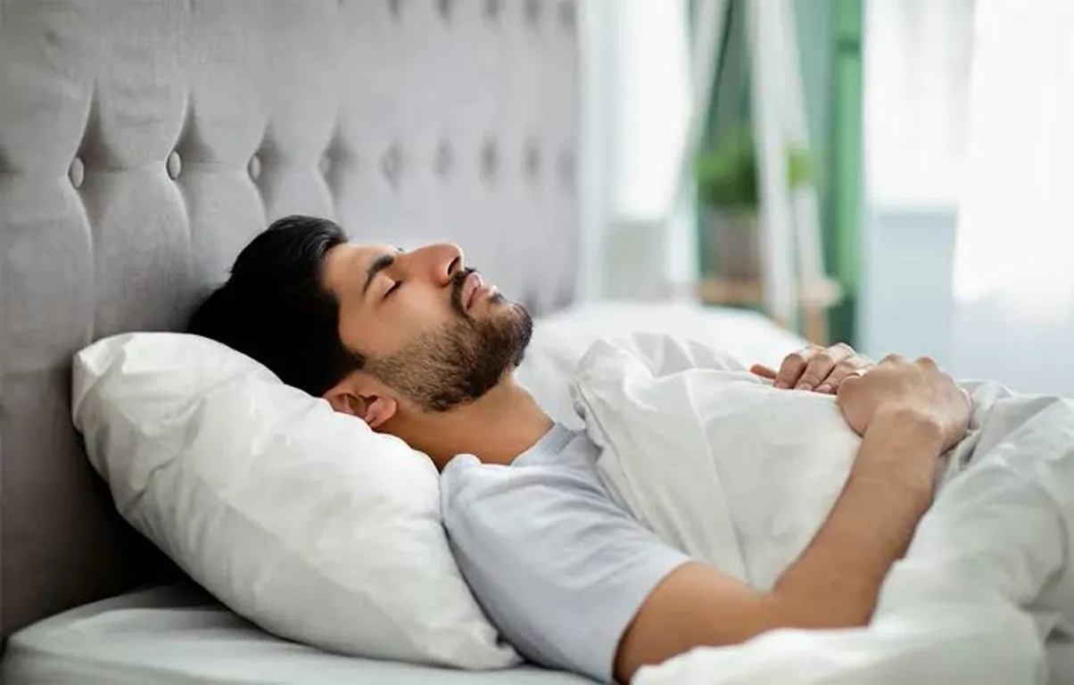 1000084736 Man sleeping peacefully in bed with white sheets, resting against a tufted headboard in a bright bedroom