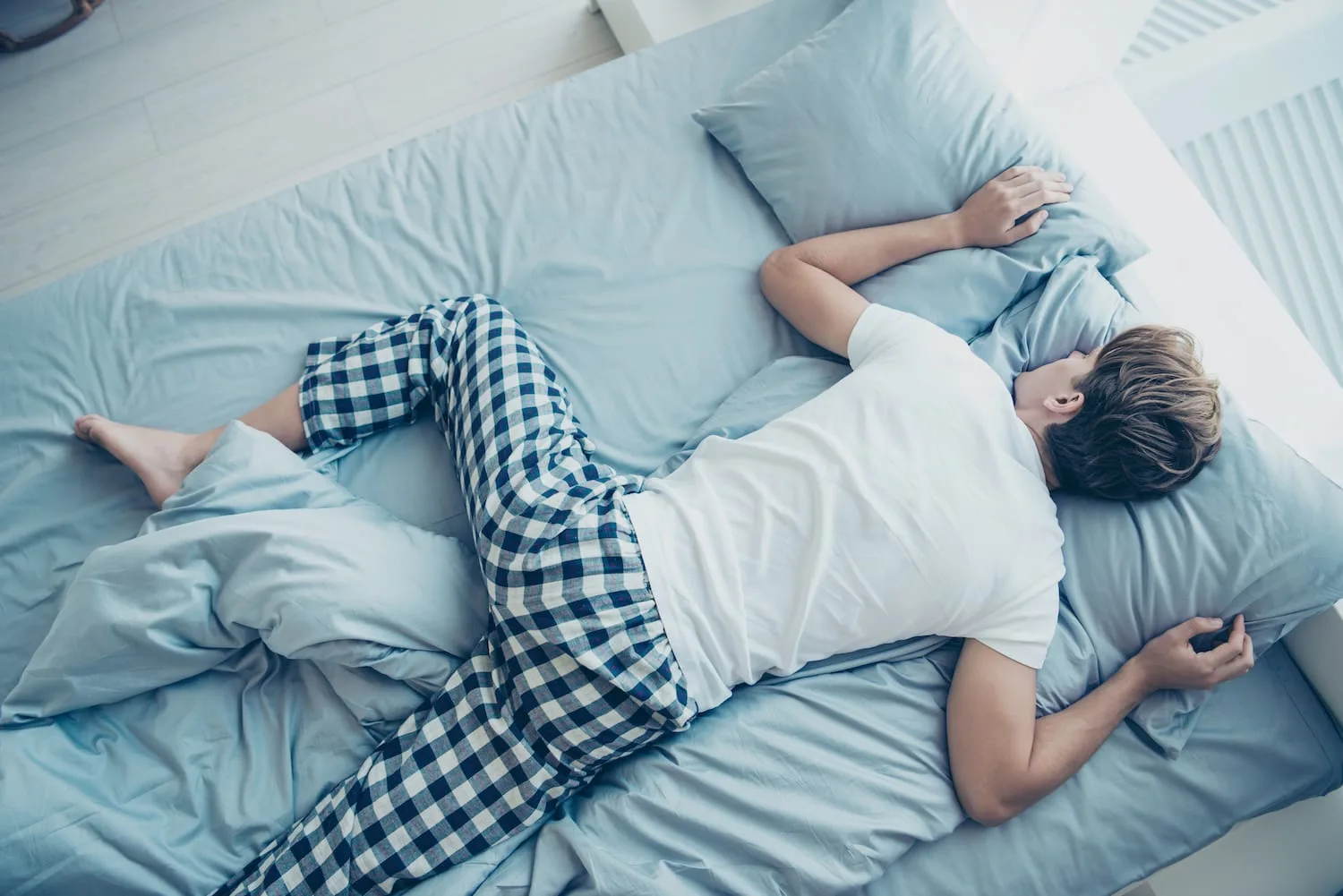 1000084727 Person lying face down on a light blue bed, wearing a white shirt and blue plaid pajama pants, resting on pillows.