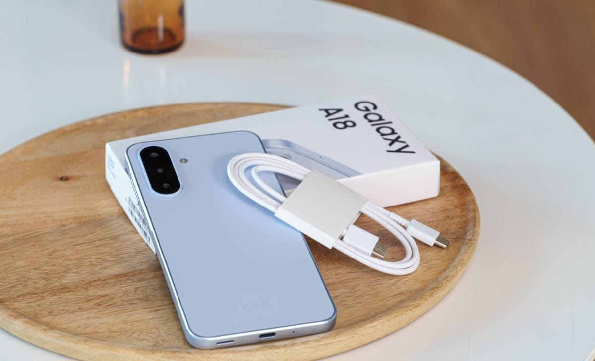 gsmarena_016 Blue Samsung Galaxy A18 smartphone with a white charging cable and adapter resting on its box on a round wooden tray.