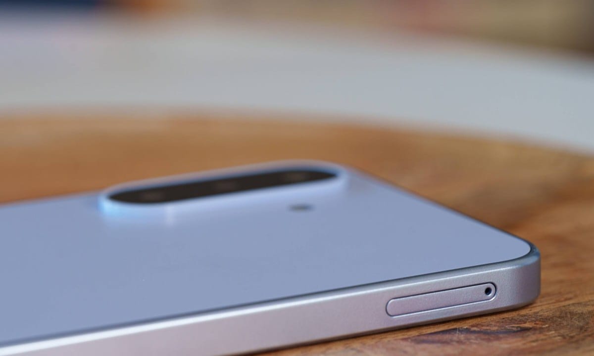 gsmarena_024 Close-up of a silver smartphone on a wooden table, highlighting its rounded dual-camera bump on the back.