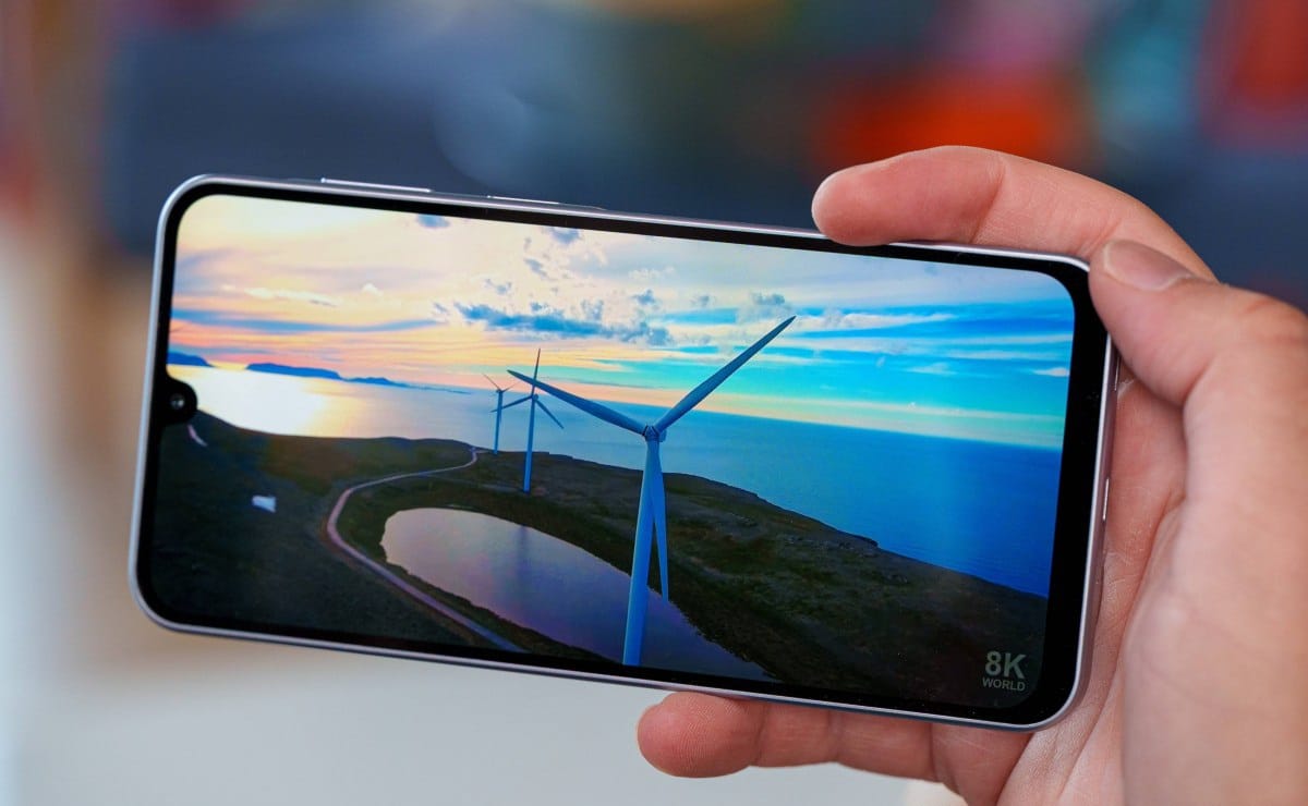 gsmarena_015 Hand holding a smartphone displaying a coastal landscape with wind turbines at sunset/deserted coast.