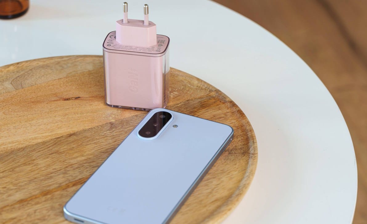 gsmarena_017 Light blue smartphone with a large camera module rests on a wooden cutting board beside a pink plug-in power adapter.